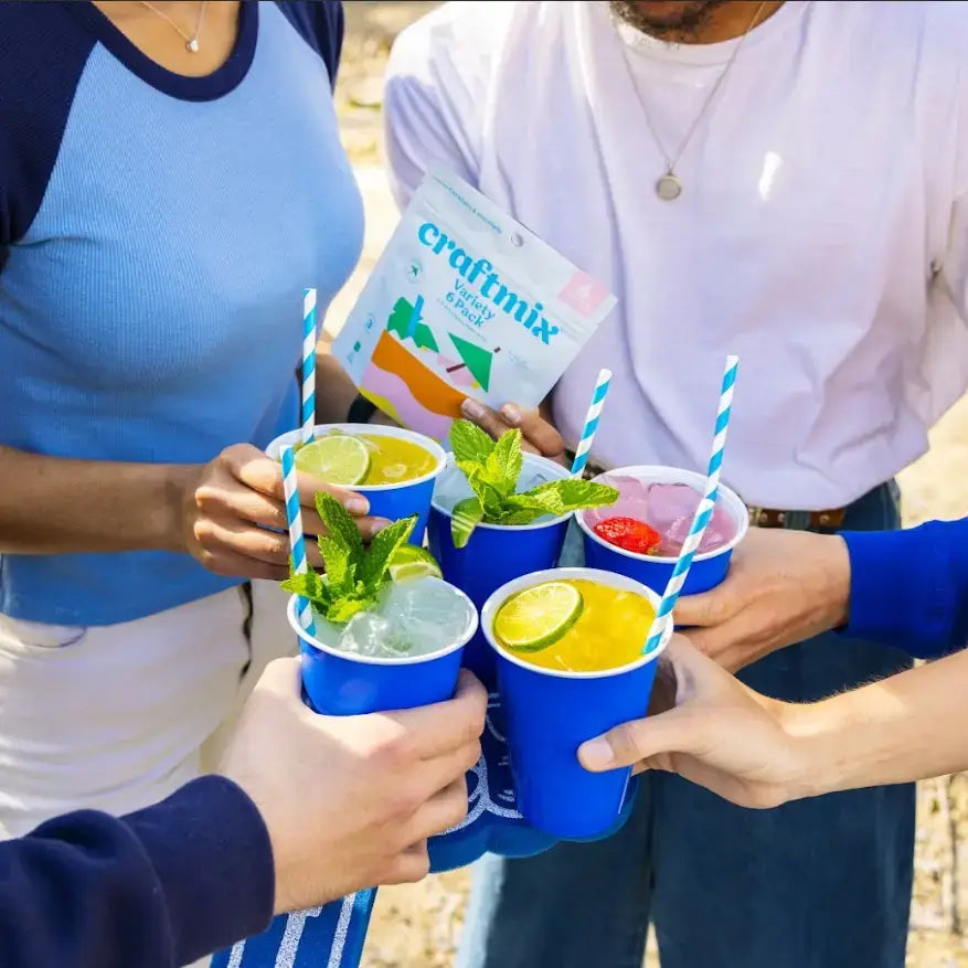 Group of friends toasting with colorful Craftmix cocktails in blue cups garnished with fresh mint and citrus